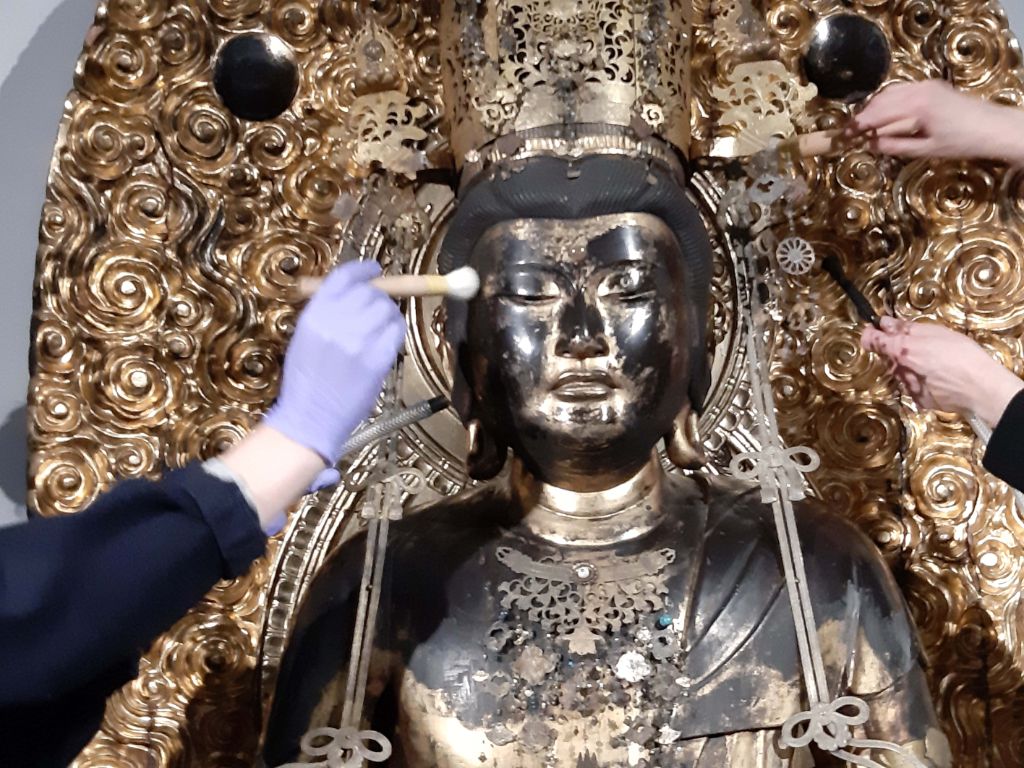 Drei Hände reinigen eine Buddhafigur, von der man Kopf und Schulterpartie sieht. Eine Hand in blauem Handschuh pinselt die rechte Schläfe des Buddha. Zwei andere Hände pinseln die Gloriole hinter dem Buddha, die aus vielen Blüten besteht.