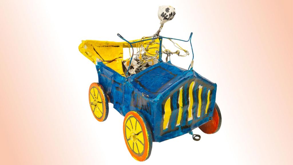 Der Weg ins Jenseits Mexikanische Totenfigur aus Pappmache, in Form eines Skeletts, welches ein blaues Oldtimer-Auto mit offenem gelben Verdeck, roten Rädern mit gelben Felgen steuert.