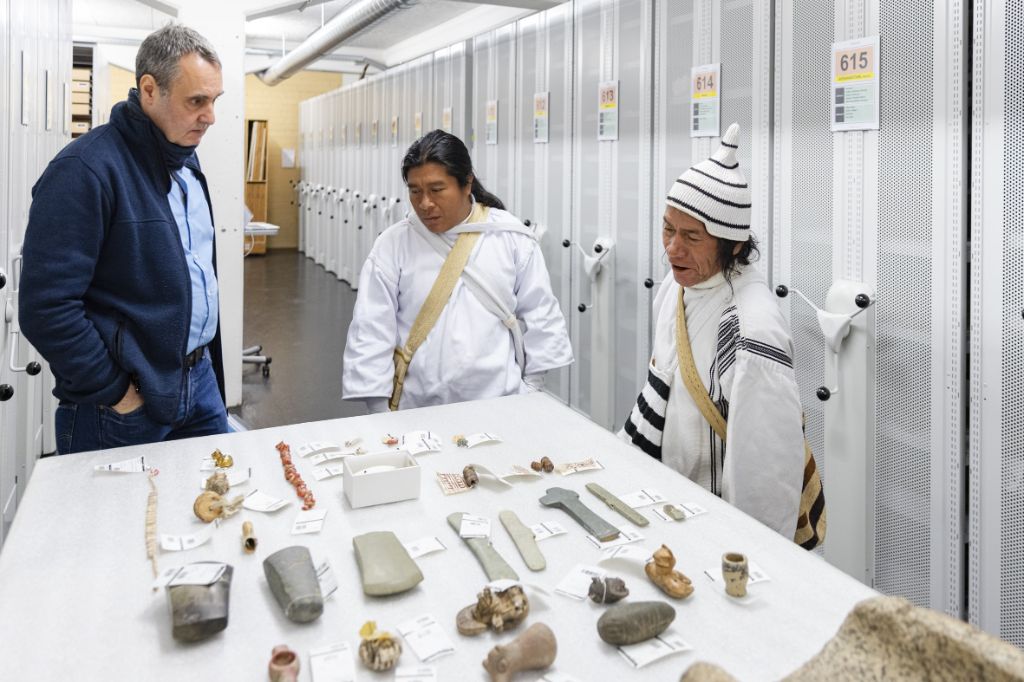 Drei Personen stehen um einen Tisch, auf dem Gegenstände mehrheitlich aus Stein liegen. Die Dinge sind mit kleinen Etiketten versehen. Der Mann rechts im Bild, gekleidet mehrheitlich in Weiss, ist am Sprechen. Die beiden anderen, ein Mann ebenfalls gekleidet in Weiss und ein Mann in in Blau hören zu. Hinter ihnen sind Rollkorpusse zu sehen.